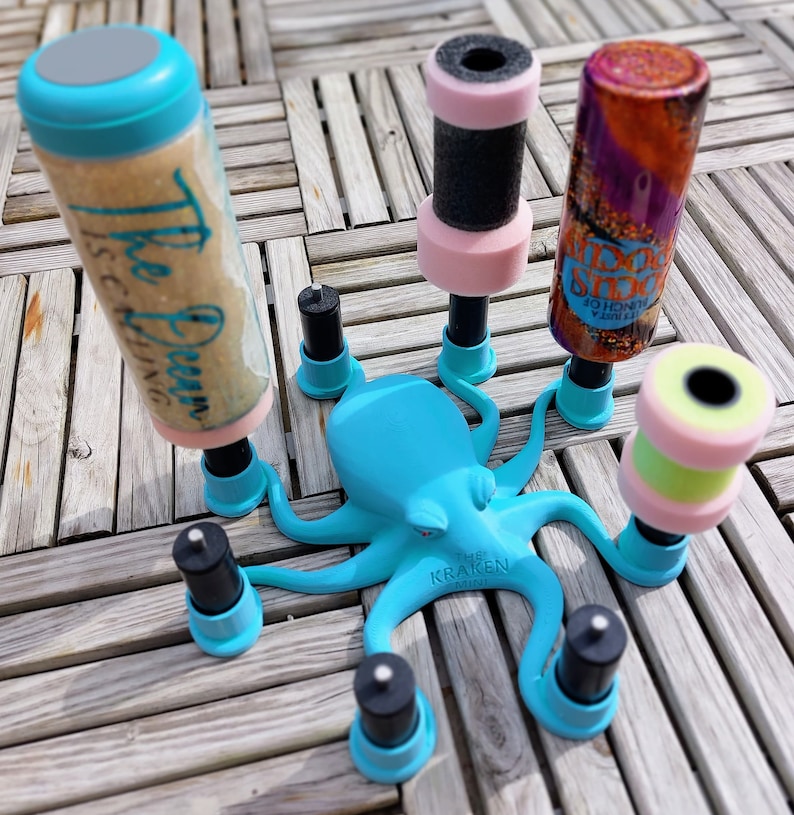 May include: A turquoise octopus-shaped holder with multiple black and pink cylindrical objects. One cylinder has the text "The Ocean is Calling". The holder is on a wooden surface. The text "The Kraken Mini" is on the octopus.