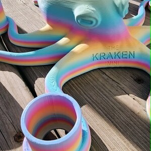 May include: A colorful, rainbow-hued octopus sculpture with the text "THE KRAKEN MINI" on its body. The sculpture is 3D-printed and features a cylindrical cup-like structure. The octopus is resting on a wooden surface.