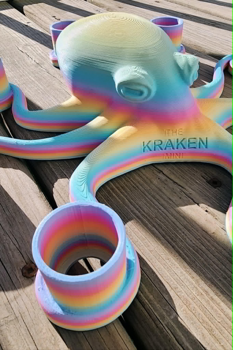 May include: A colorful, rainbow-hued octopus sculpture with the text "THE KRAKEN MINI" on its body. The sculpture is 3D-printed and features a cylindrical cup-like structure. The octopus is resting on a wooden surface.