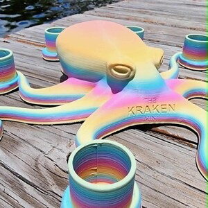 May include: A rainbow-colored octopus sculpture with the text "THE KRAKEN MINI" on its side. The octopus is surrounded by several matching rainbow-colored cylindrical objects. The items are on a wooden dock near water.