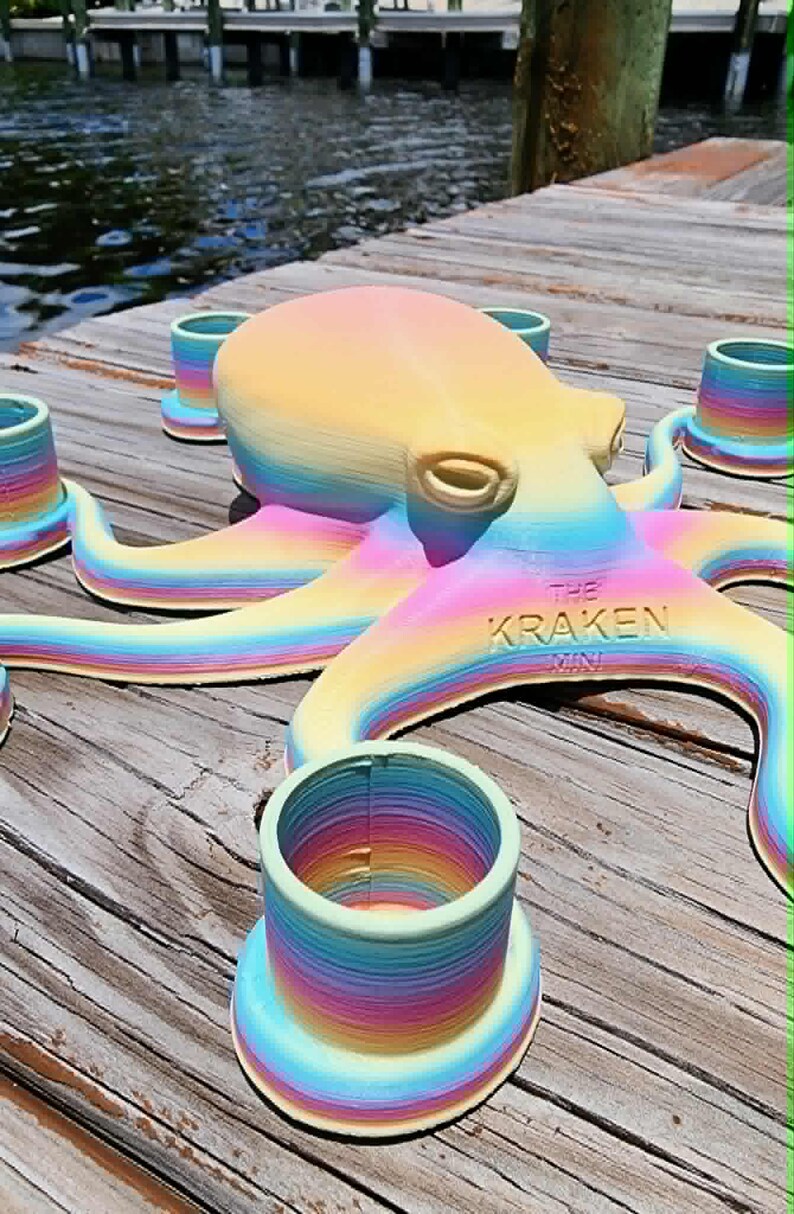 May include: A rainbow-colored octopus sculpture with the text "THE KRAKEN MINI" on its side. The octopus is surrounded by several matching rainbow-colored cylindrical objects. The items are on a wooden dock near water.