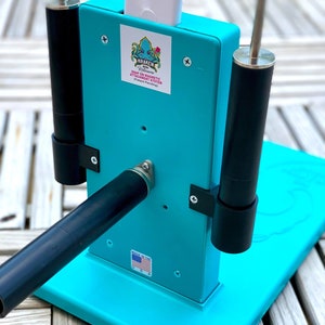 Op de afbeelding: Een turquoise blauwe metalen standaard met een zwarte en zilveren stang en twee zwarte palen. De standaard heeft een wit apparaat met een zwarte knop en een sticker met de tekst "Kraken Robotics Snap-On Magnetic Attachment System (Patent Pending)". De standaard heeft ook een sticker met de tekst "Made in the USA".