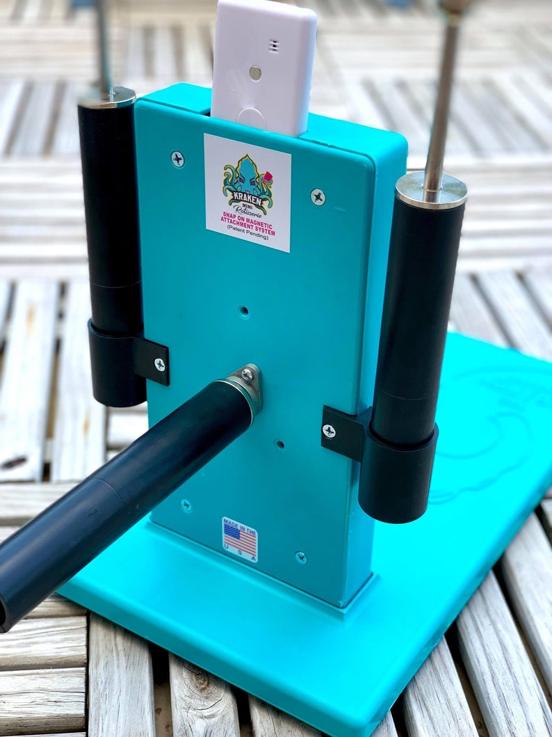 Op de afbeelding: Een turquoise blauwe metalen standaard met een zwarte en zilveren stang en twee zwarte palen. De standaard heeft een wit apparaat met een zwarte knop en een sticker met de tekst "Kraken Robotics Snap-On Magnetic Attachment System (Patent Pending)". De standaard heeft ook een sticker met de tekst "Made in the USA".