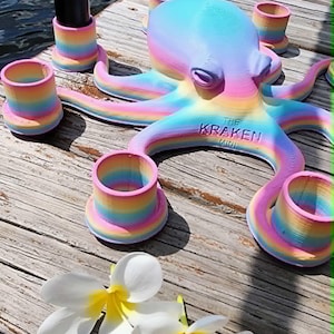 May include: A colorful octopus sculpture with the text "THE KRAKEN MINI" is surrounded by matching rainbow-colored candle holders. Two white flowers with yellow centers are in the foreground. The octopus and candle holders are on a wooden surface.