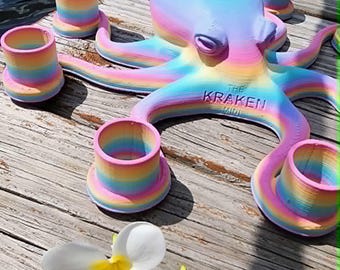 May include: A colorful octopus sculpture with the text "THE KRAKEN MINI" is surrounded by matching rainbow-colored candle holders. Two white flowers with yellow centers are in the foreground. The octopus and candle holders are on a wooden surface.