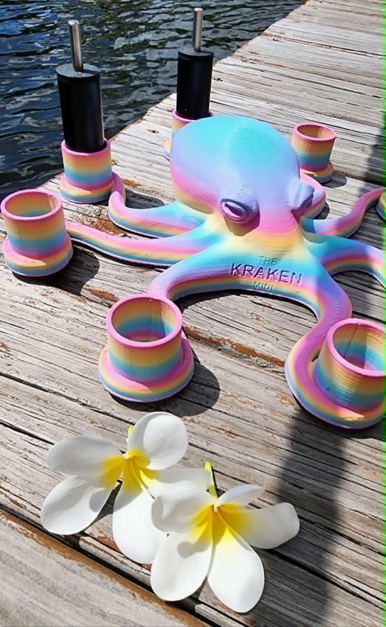 May include: A colorful octopus sculpture with the text "THE KRAKEN MINI" is surrounded by matching rainbow-colored candle holders. Two white flowers with yellow centers are in the foreground. The octopus and candle holders are on a wooden surface.