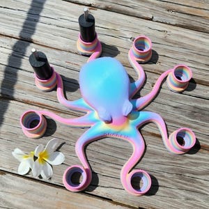 May include: A colorful octopus-shaped nail polish holder in pastel shades of pink, blue, and yellow. The octopus has eight arms with circular holders and two black nail polish bottles. A white flower is next to the octopus.