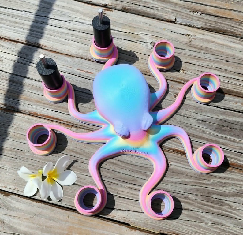 May include: A colorful octopus-shaped nail polish holder in pastel shades of pink, blue, and yellow. The octopus has eight arms with circular holders and two black nail polish bottles. A white flower is next to the octopus.
