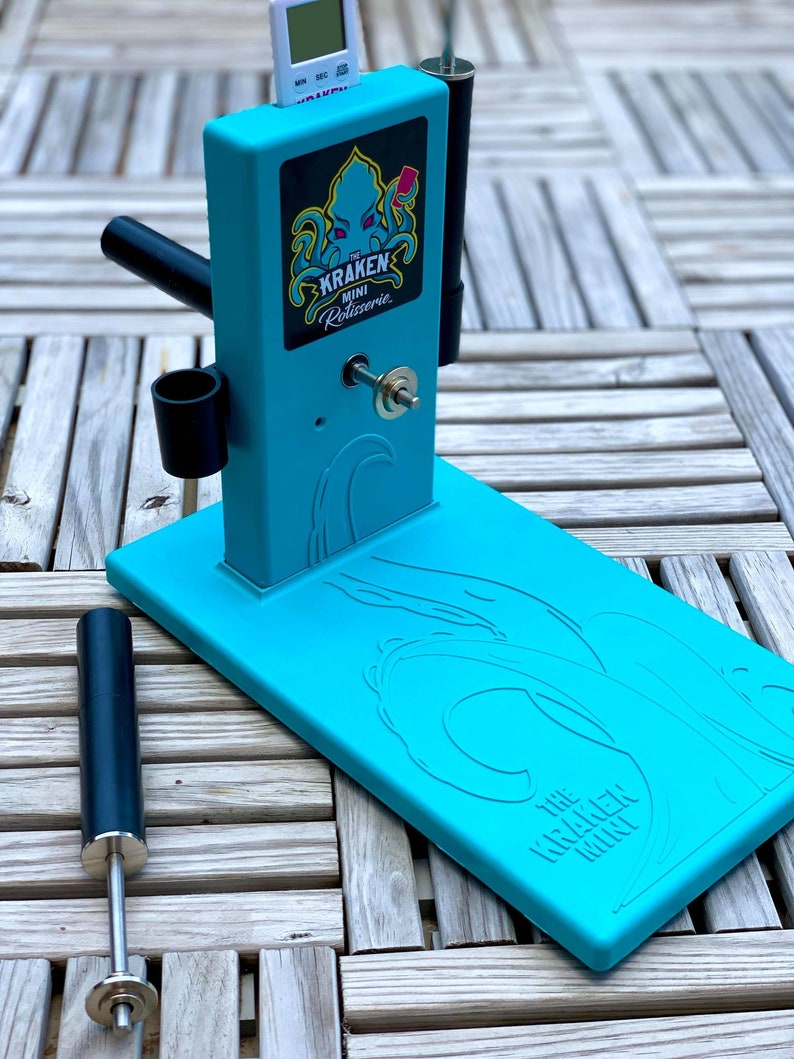 Op de afbeelding: Een turquoise blauwe rotisserie met een zwarte handgreep en een digitale timer. De rotisserie heeft een zwart-wit logo met de tekst "The Kraken Mini Rotisserie".