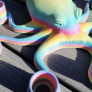 May include: A rainbow-colored octopus sculpture with the text "THE KRAKEN MINI" on its underside. The sculpture has multiple tentacles and is surrounded by matching ring-shaped objects. The octopus is made of a material that appears to be 3D-printed.
