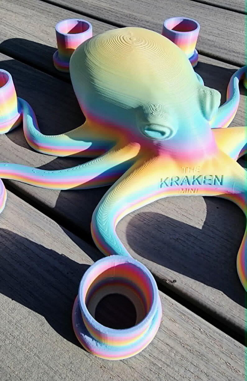 May include: A rainbow-colored octopus sculpture with the text "THE KRAKEN MINI" on its underside. The sculpture has multiple tentacles and is surrounded by matching ring-shaped objects. The octopus is made of a material that appears to be 3D-printed.