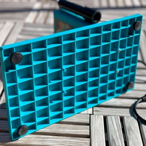Op de afbeelding: De onderkant van een turquoise blauwe metaaldetector met een rasterpatroon en vier zwarte rubberen voetjes. De detector is aangesloten op een stopcontact.