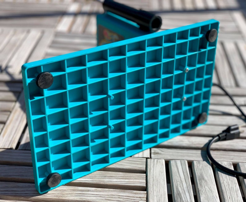 Op de afbeelding: De onderkant van een turquoise blauwe metaaldetector met een rasterpatroon en vier zwarte rubberen voetjes. De detector is aangesloten op een stopcontact.