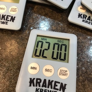 May include: A white digital timer with a black display showing 02:00. The timer has buttons for minutes, seconds, and stop/start. The timer is labeled "Kraken Krewe".