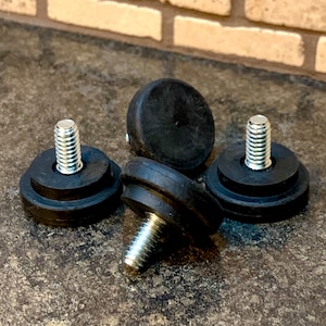 May include: Four black rubber furniture feet with silver threaded bolts. The feet have a circular design with a flat base and a rounded top. The bolts are centered in the feet and are designed for screwing into furniture legs.