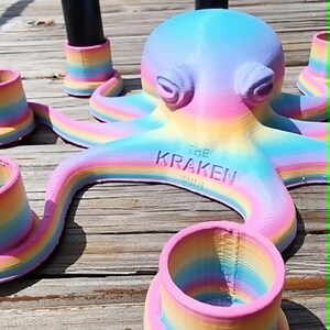 May include: A 3D-printed octopus sculpture in pastel rainbow colors, with the text "THE KRAKEN" on its body. The sculpture is surrounded by matching rainbow-colored cup-shaped objects and black cylindrical objects with silver tops.