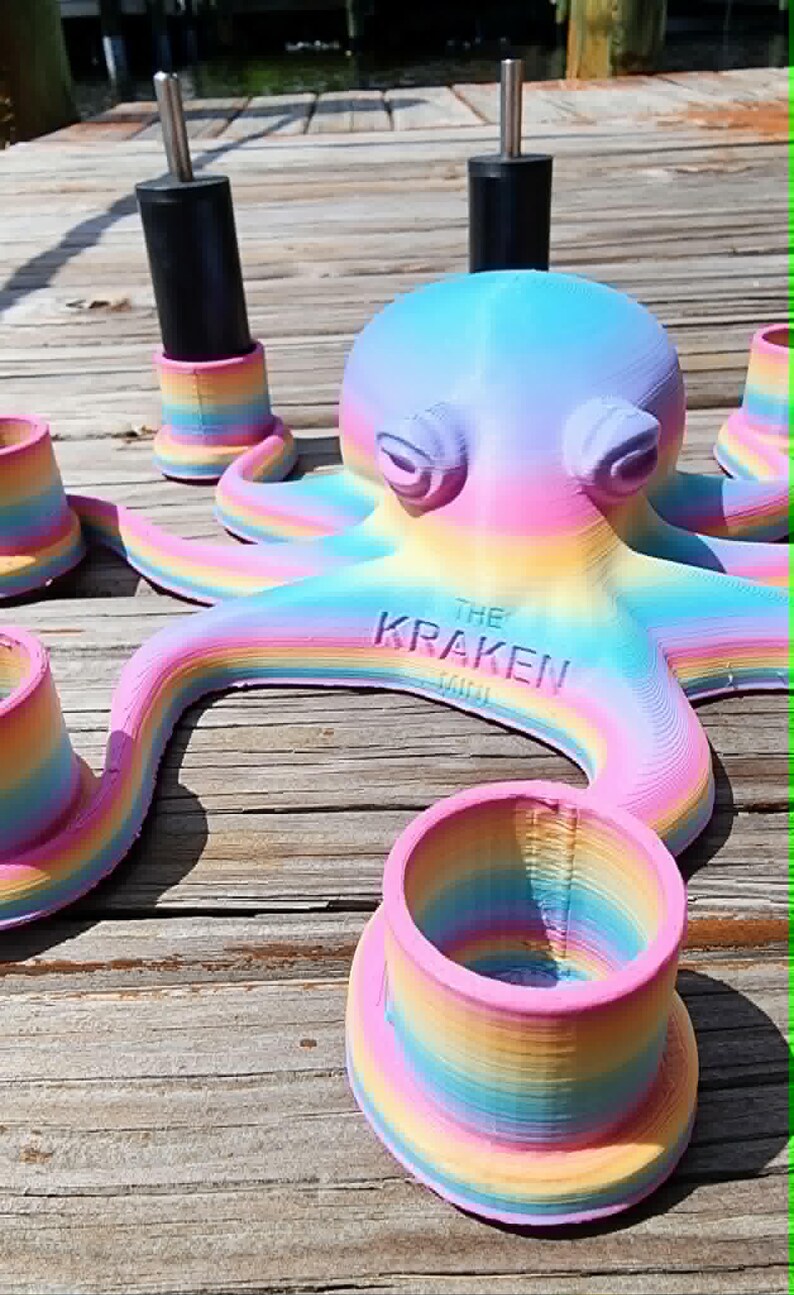 May include: A 3D-printed octopus sculpture in pastel rainbow colors, with the text "THE KRAKEN" on its body. The sculpture is surrounded by matching rainbow-colored cup-shaped objects and black cylindrical objects with silver tops.