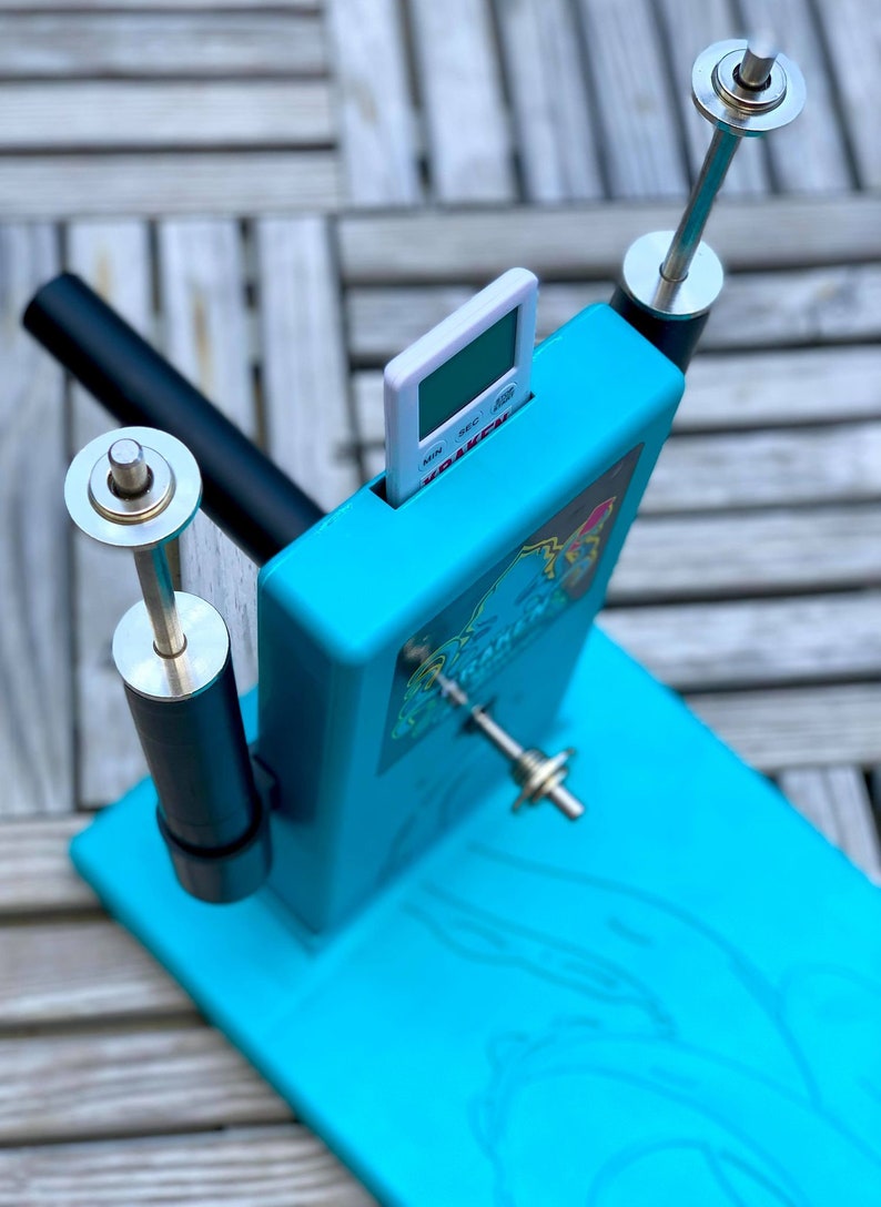 Op de afbeelding: Een turquoise blauw plastic apparaat met een witte digitale timer, twee zwarte en zilveren staven en een zilveren draaiende staaf. Het apparaat heeft een afbeelding van een cartoonfiguur aan de voorkant.