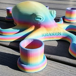 May include: A colorful octopus-shaped shot glass holder with eight matching shot glasses. The octopus and glasses are rainbow-colored, with the text "THE KRAKEN MINI" on the octopus. The set is displayed on a wooden surface.