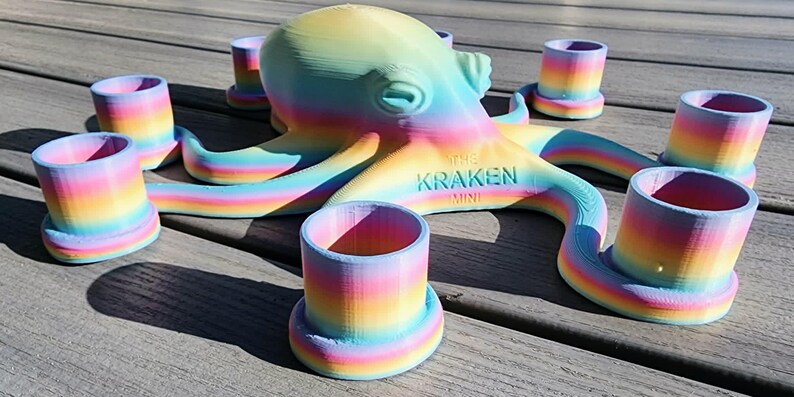 May include: A colorful octopus-shaped shot glass holder with eight matching shot glasses. The octopus and glasses are rainbow-colored, with the text "THE KRAKEN MINI" on the octopus. The set is displayed on a wooden surface.