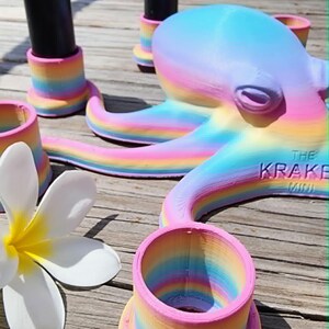 May include: A whimsical, rainbow-colored octopus sculpture with the text "THE KRAKE MINI" on its side. The octopus is surrounded by matching rainbow-striped holders and a white flower with a yellow center. The items are on a wooden surface.