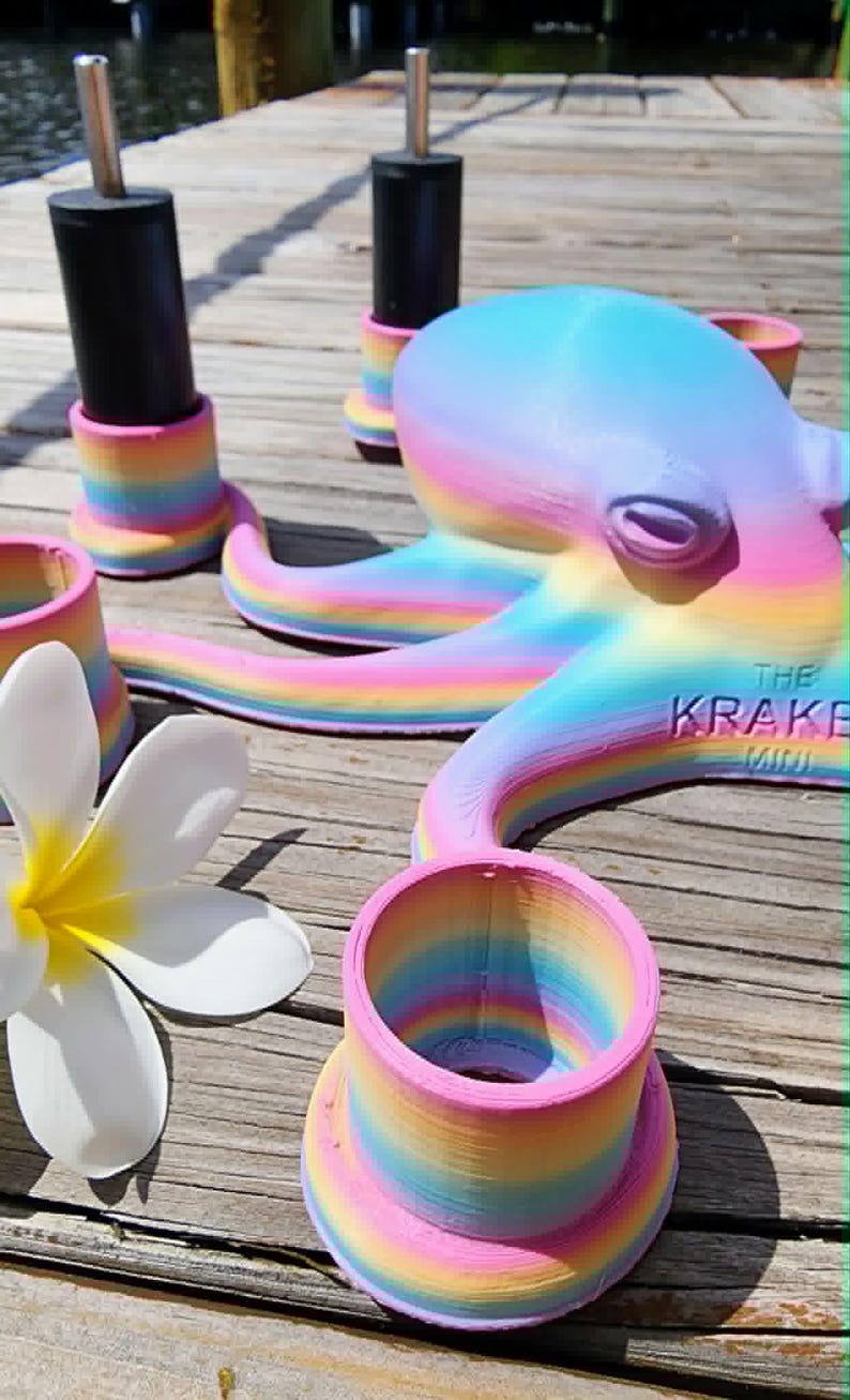 May include: A whimsical, rainbow-colored octopus sculpture with the text "THE KRAKE MINI" on its side. The octopus is surrounded by matching rainbow-striped holders and a white flower with a yellow center. The items are on a wooden surface.