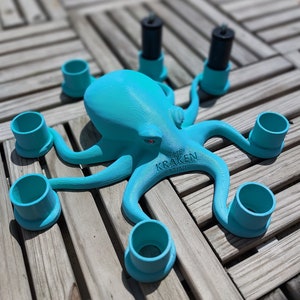 May include: A turquoise octopus-shaped candle holder with six arms, each holding a teal cylinder. Two black candles are in the center. The text "THE KRAKEN MINI" is on the base. The octopus has red eyes.