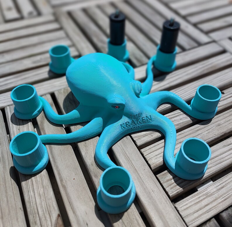 May include: A turquoise octopus-shaped candle holder with six arms, each holding a teal cylinder. Two black candles are in the center. The text "THE KRAKEN MINI" is on the base. The octopus has red eyes.