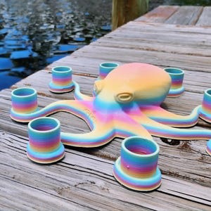 May include: A colorful octopus-shaped candle holder with seven cylindrical candle cups. The octopus and candle cups are painted in a rainbow gradient of colors. The candle holder is sitting on a wooden dock near water.