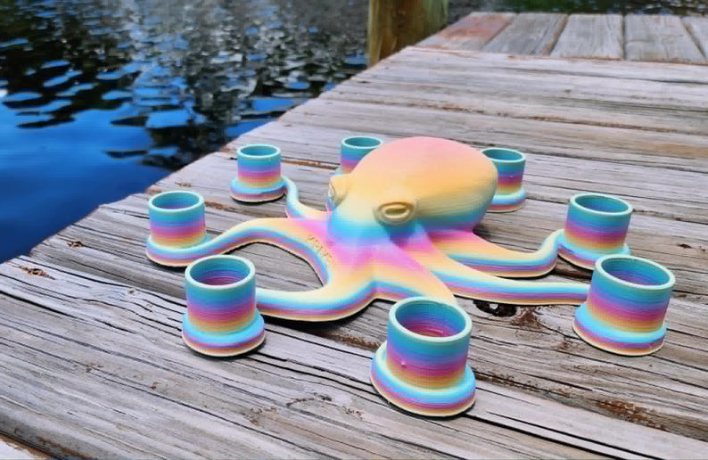 May include: A colorful octopus-shaped candle holder with seven cylindrical candle cups. The octopus and candle cups are painted in a rainbow gradient of colors. The candle holder is sitting on a wooden dock near water.