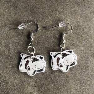 Peut inclure: Une paire de boucles d'oreilles argent&eacute;es avec un charme de vache de dessin anim&eacute; blanc et noir. Chaque vache tient une petite bouteille de lait.