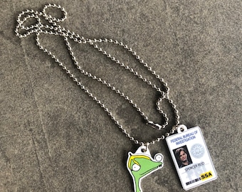 May include: A silver chain necklace with two pendants. One pendant is a cartoon drawing of a green creature with a yellow head. The other pendant is a fake ID card with the text "Federal Bureau of Investigation" and "Spencer Reid" printed on it.