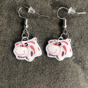 Peut inclure: Une paire de boucles d'oreilles pendantes argent&eacute;es repr&eacute;sentant une vache de dessin anim&eacute; avec des taches roses et blanches. La vache sourit et a une fraise rouge dans la bouche.