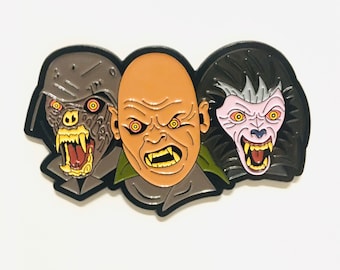 Op de afbeelding: Een zwarte emaille pin met drie monstergezichten. De gezichten zijn een weerwolf, een wezen met gloeiende rode ogen en een kale man met gloeiende rode ogen. Alle drie de gezichten grommen met scherpe tanden.