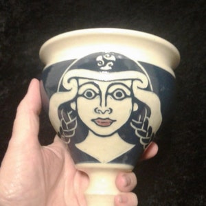 May include: A ceramic goblet with a blue and white design. The goblet features a stylized image of a woman's face with a crown on her head.