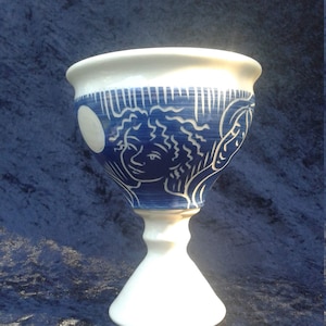 Könnte beinhalten: Ein weißer Keramikkelch mit einem blauen und weißen Design. Das Design zeigt ein stilisiertes Gesicht und einen Mond.