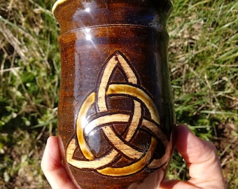 May include: A brown ceramic mug with a Celtic knot design in a lighter brown color.