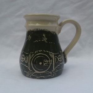 Peut inclure: Une tasse en céramique avec un motif vert et blanc représentant une porte ronde avec un cadre blanc et un cercle blanc au centre. La tasse a une poignée blanche et un bord blanc.
