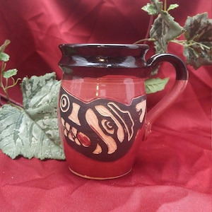 May include: A red ceramic mug with a black rim and handle. The mug has a stylized black and white design of an animal's face on the front.