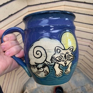 May include: A blue ceramic mug with a white raccoon design and a yellow moon. The raccoon is facing the viewer and has a swirl pattern on its tail.