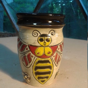 May include: A ceramic mug with a hand-painted design of a bee with a human face. The bee is yellow and black with red wings and a white body. The mug has a black rim and a white background.