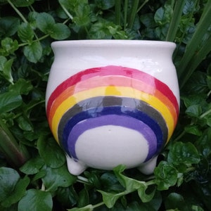 Puede incluir: Maceta de cerámica blanca con tres patas y un diseño de arcoíris pintado en la parte delantera. El arcoíris tiene rayas rojas, naranjas, amarillas, verdes, azules, índigo y violetas.