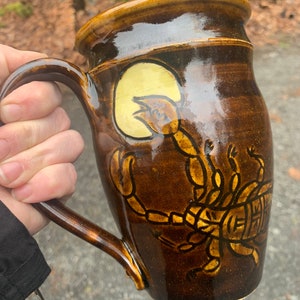 Può includere: Una tazza in ceramica marrone con manico. La tazza ha un design di scorpione giallo e marrone sul davanti.