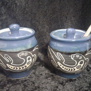 May include: Two blue ceramic honey pots with lids and a white design of an otter on each side. One pot has a wooden honey dipper.