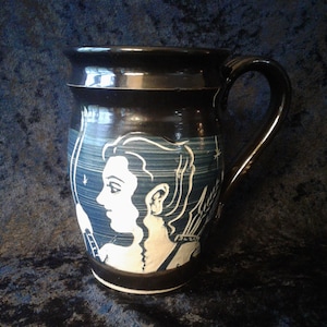 May include: A black ceramic mug with a white design of a woman's face with a wing on her shoulder. The mug has a handle and is decorated with a white design.