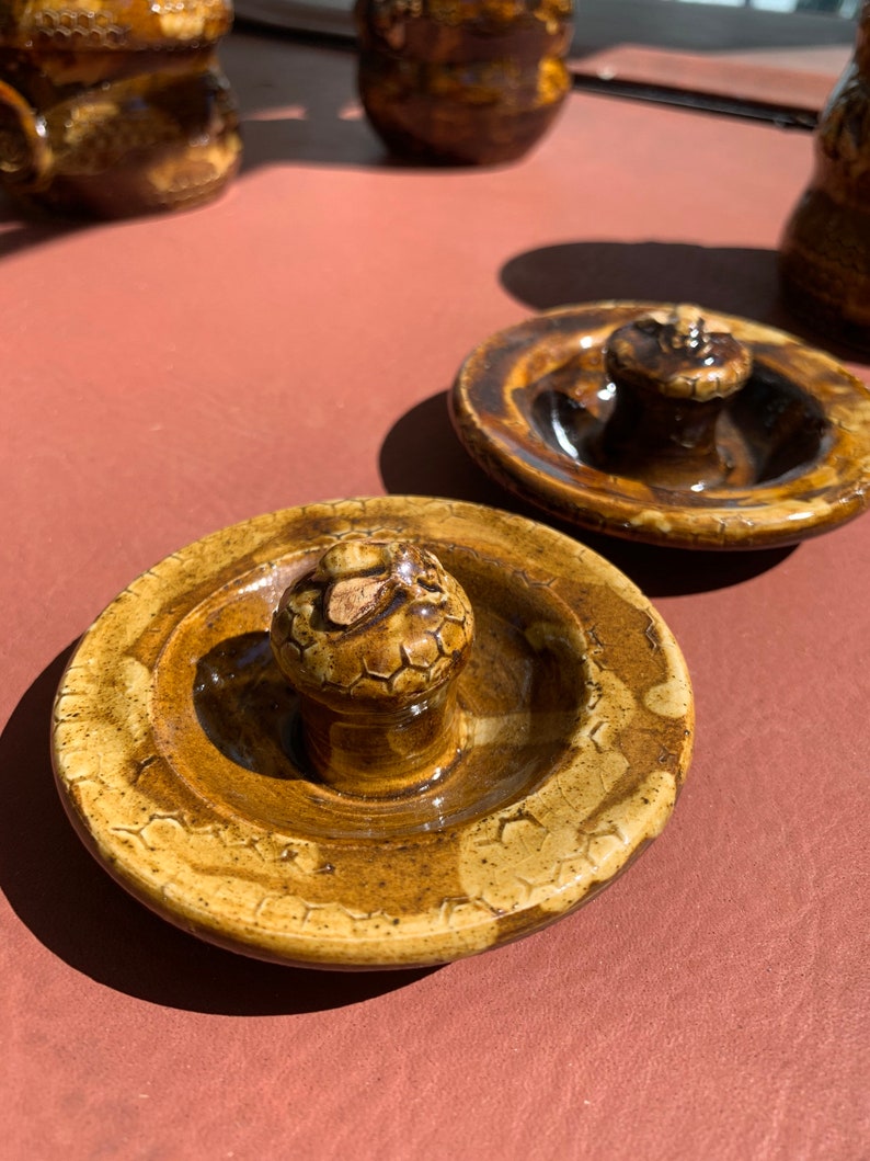 Op de afbeelding: Twee bruine keramische deksels met een verhoogde knop in het midden. De deksels hebben een getextureerd oppervlak met een honingraatpatroon.