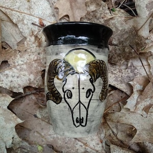 May include: A ceramic mug with a black rim and a white background. The mug features a black and brown illustration of a ram skull with horns.