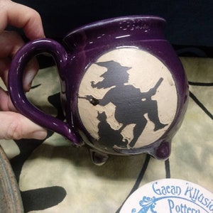 May include: Purple ceramic mug with a white silhouette of a witch flying on a broomstick with a black cat. The mug has three feet and a handle.