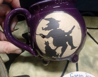 May include: Purple ceramic mug with a white silhouette of a witch flying on a broomstick with a black cat. The mug has three feet and a handle.