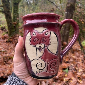 May include: A burgundy ceramic mug featuring a white fox design. The fox is curled up with its tail wrapped around its body. The mug has a handle and is ideal for enjoying a hot drink.
